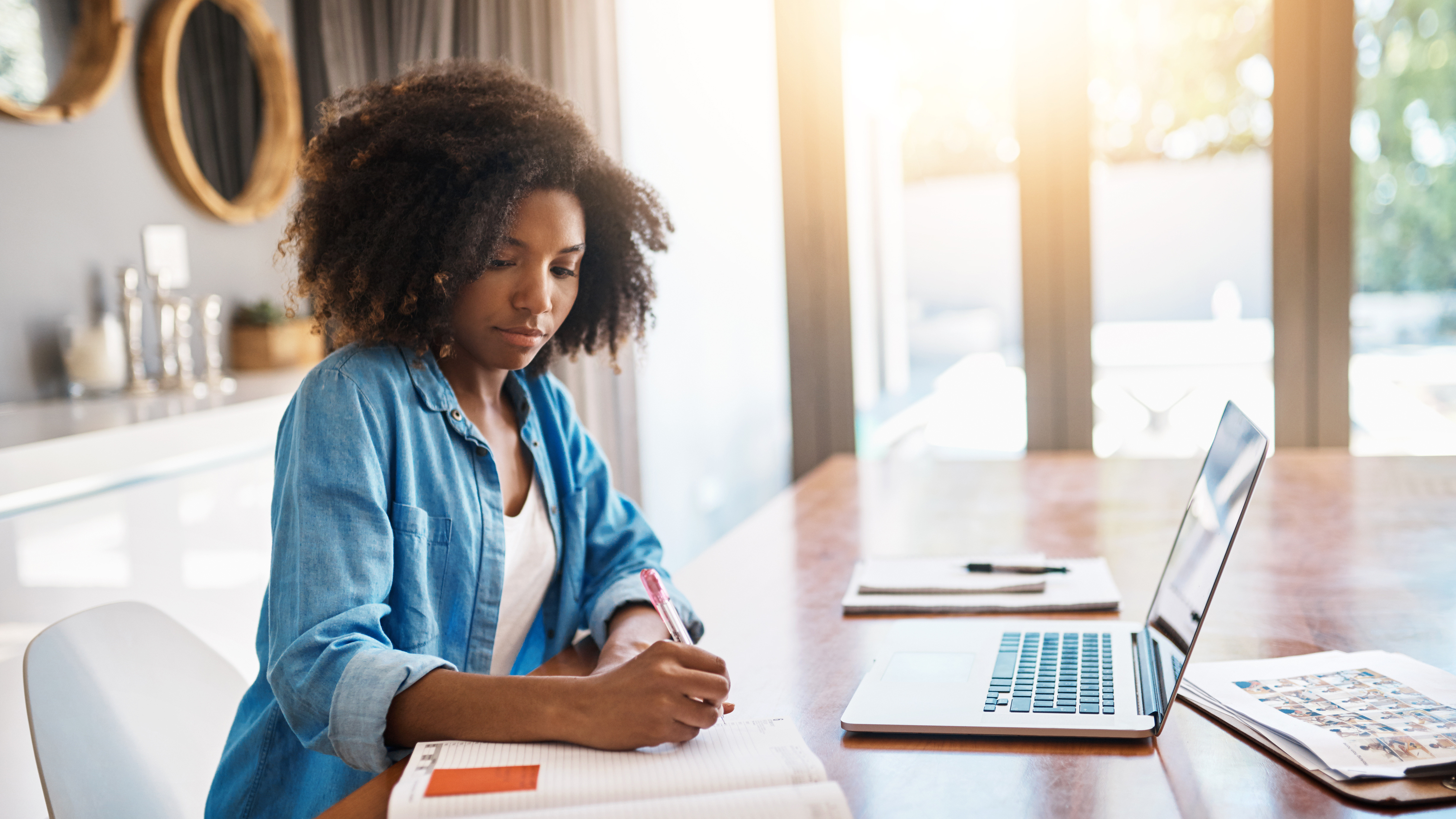 A student sitting at a table with a laptop in front of her and writing in a notebook